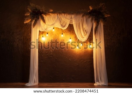 arch with white fabric against a brown wall with flowers. wedding classic arch for a wedding photo shoot. wooden arch with dry flowers and incandescent lamps in a photo studio Royalty-Free Stock Photo #2208474451