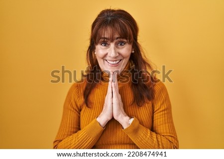 Middle age hispanic woman standing over yellow background praying with hands together asking for forgiveness smiling confident. 