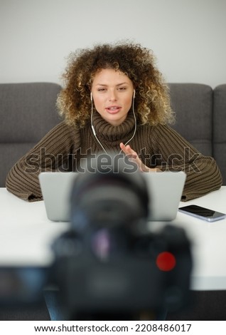 Video blogger woman speaking on camera. Cute curly female in headset talking on videocamera. Download stock photo of girl filming educational videoblog