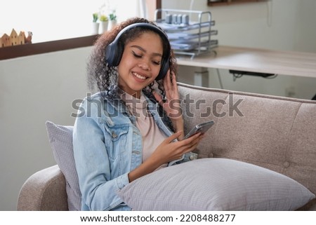 An African American woman uses a smartphone and puts on a music headset in her home to relax.
