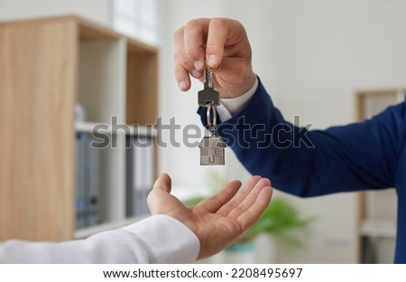 Professional realtor gives buyer key to new home. Real estate agent hands over key to young homeowner. Cropped close up shot male hand holding key with house shaped pendant. Property purchase concept Royalty-Free Stock Photo #2208495697