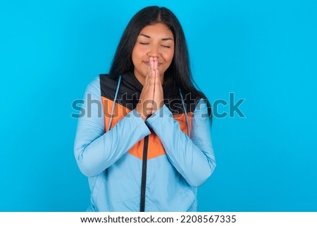 Indoor closeup of Young latin woman wearing sport clothes over blue background practicing yoga and meditation, holding palms together in namaste, looking calm, relaxed and peaceful.