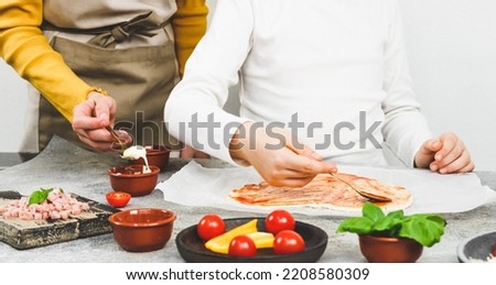 Two pairs of hands of Caucasian girls sisters smear sauce on spoons on pizza heart dough with ingredients on the table, side view close-up. Valentine's day cooking pizza concept.