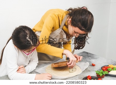 Caucasian girls sisters together cuts dough on a wooden plate with a metal scabbard with ingredients on the table, close-up side view. Concept of cooking pizza for valentine's day.