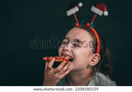 Happy little caucasian girl with closed eyes and a rim on her head made of santa claus hats holds in her hand a slice of pizza near her mouth on the right on a black background with copy space on the 