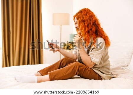 Adolescent teen girl texting on a smartphone sitting in bed at home in her bedroom with a cup of coffe.