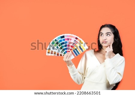 Beautiful brunette woman with thoughtful face holding color palette fan on orange background with copy space.