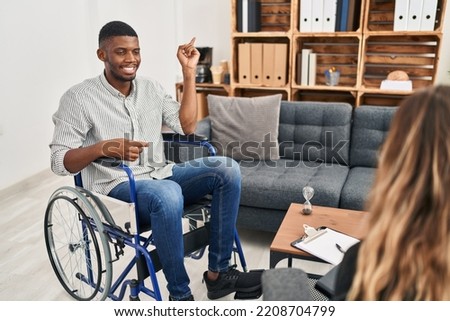African american man doing therapy sitting on wheelchair with a big smile on face, pointing with hand and finger to the side looking at the camera. 