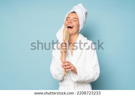 Funny portrait of happy woman in bathrobe and towel on head standing on blue studio background with closed eyes enjoying singing in massage brush as in microphone pretending to be star singer Royalty-Free Stock Photo #2208723233