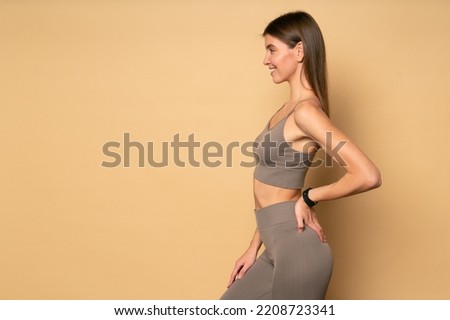 Profile view of sporty trainer in crop top and leggings standing with hand on hip on light brown background with copy space on the right, showing exercises, wearing fitness tracker on wrist Royalty-Free Stock Photo #2208723341