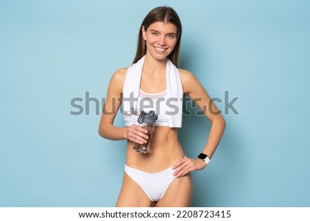 Portrait of smiling sporty woman in stylish sports bikini wearing fitness tracker having break in workout session with towel on shoulders, holding bottle of water on blue studio background