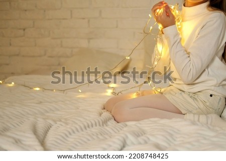 A girl in a white sweater and skirt sits on the bed and holds a Christmas garland in her hands. Horizontal photo