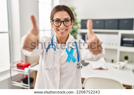 Young brunette doctor woman wearing stethoscope at the clinic approving doing positive gesture with hand, thumbs up smiling and happy for success. winner gesture. 
