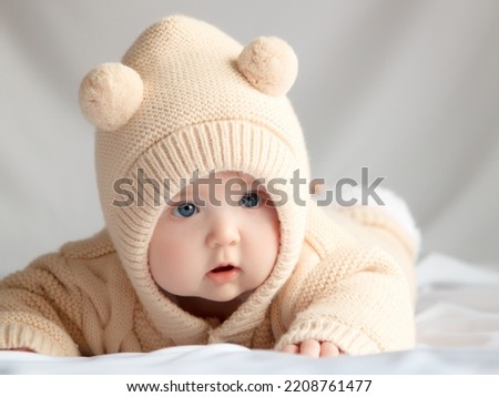 Just beautiful. Cute smiling baby. Cute 3 month old Baby girl infant on a bed on her belly with head up looking with her big eyes. Warm, fluffy biege clothes. Closeup. Three months old baby.