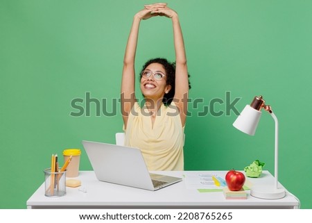 Young employee business woman of African American ethnicity wear shirt sit work at white office desk with pc laptop stretch hands rest isolated on plain green background. Achievement career concept. Royalty-Free Stock Photo #2208765265