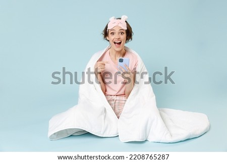 Full length young shock woman in pajamas jam sleep eye mask rest at home sit wrap cover blanket duvet point finger mobile cell phone isolated on pastel blue background Good mood night bedtime concept Royalty-Free Stock Photo #2208765287