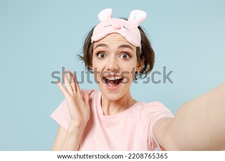 Close up young fun woman in pajamas jam sleep eye mask rest relaxing at home do selfie shot mobile phone spread hand isolated on pastel blue background studio portrait. Good mood night bedtime concept