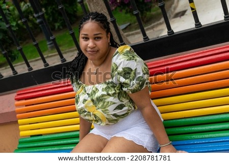 Black woman sitting on a bench painted with the LGBTI flag. Young plus size girl sitting on a bench in the street while she poses.