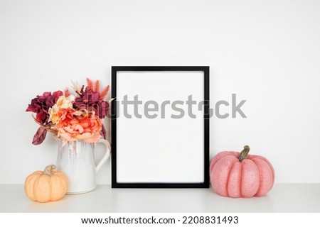 Mock up black picture frame with pink hue autumn flowers and pumpkins on a white shelf against a white wall. Fall concept. Copy space.
