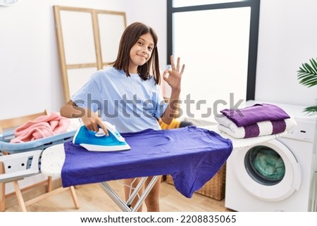 Young hispanic girl ironing clothes at laundry room doing ok sign with fingers, smiling friendly gesturing excellent symbol 