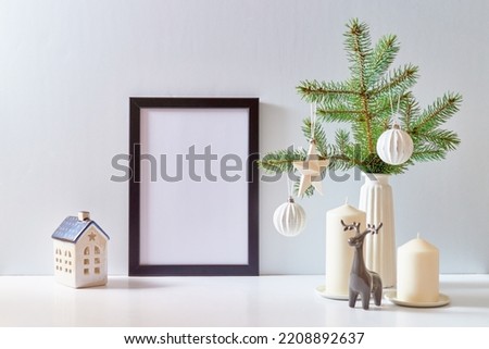 Mock up black poster frame with christmas decoration in home interior, scandinavian style. Green fir branches in a vase, deer and ball on a white table