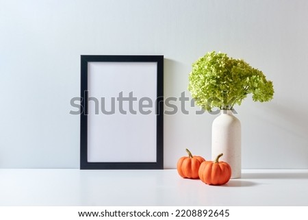 Mockup with a black frame and green flowers in a vase, pumpkins on a light background. Empty poster frame mockup for presentation design, text, lettering