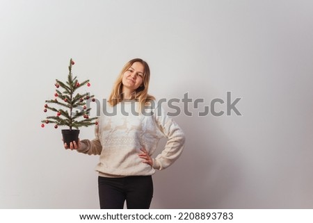 girl in a knitted sweater with deer and a Christmas tree in her hand. Slav girl in a knitted New Year's pullover with animals on a light wall. Christmas sweater on a woman for a New Year's photo shoot