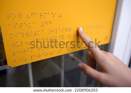 Girl reads a yellow Braille sign outdoors. Close-up of a woman's hand reading Braille. The hand of a child  touches the bulges of the plate. World Sight Day. World Braille Day. 