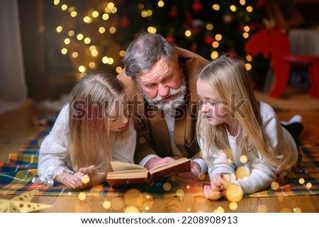 An elderly man with a beard reads a book on the floor to his granddaughters against the backdrop of a Christmas tree. 