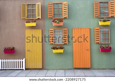 Colorful wooden yellow and orange door with windows on green and brown wall, white fence and flowers in pots, shutters open. decorative vintage house concept Royalty-Free Stock Photo #2208927175