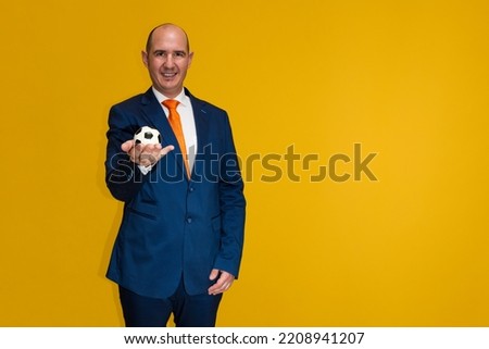 A bald Caucasian bald adult male wearing a blue suit, white shirt and orange tie holds a small soccer ball in his hand while looking at the camera. The background is yellow.