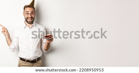 Holidays and celebration. Happy man enjoying birthday party, holding bday cake and pointing finger left at promo, standing over white background