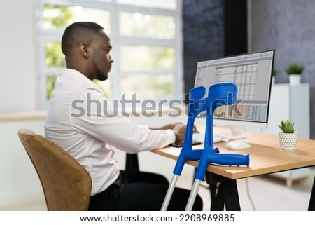 Worker With Crutches At Workplace. Social Security Compensation Royalty-Free Stock Photo #2208969885