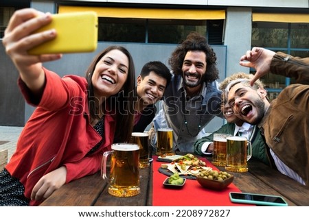 Happy multiracial group of friends enjoying beer and food together take selfie with cell phone in a bar outdoors.
