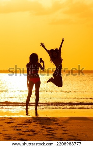 Silhouette of young female taking photo of friend jumping by tropical ocean at sunset with orange sky Bahamas