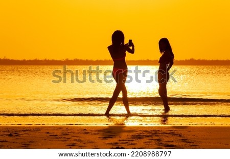 Silhouette of girls in swimwear using phone taking photos in tropical ocean at sunrise with orange sky Bahamas