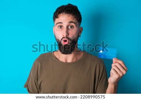 Photo of young hispanic bearded man standing against blue background amazed shocked hold credit card payment