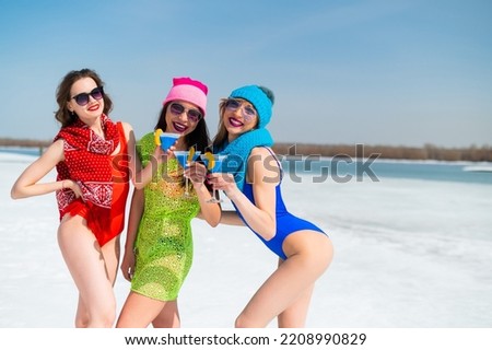 Three female friends in swimsuits drink blue alcoholic cocktails on a snowy beach. Hot girls posing in bikinis outdoors in winter.