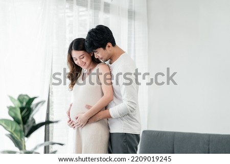 An asian couple seems very happy. A handsome husband is  embracing his beautiful pregnant wife next to the window in the living room. Royalty-Free Stock Photo #2209019245