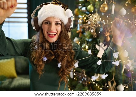 Christmas time. smiling trendy 40 years old woman with Santa hat in green dress listening to the music with headphones and dancing near Christmas tree in the modern house.
