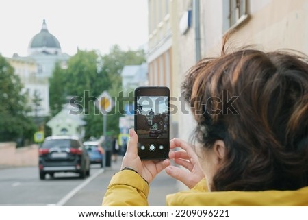 Elderly brunette woman takes pictures of Orthodox church on smartphone. Tourist woman photographs landmark while walking around the city. Focus on smartphone screen, blurred foreground and background