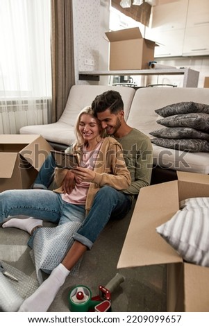 Young happy multiethnic couple resting and looking photo on floor during packing things in cardboard box. Middle eastern man and blonde caucasian woman moving to new apartment. Home relocation