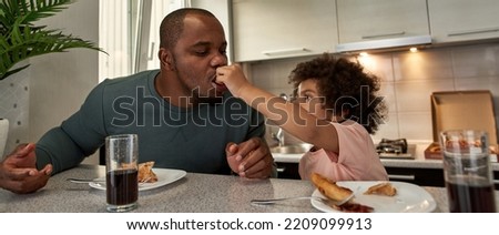 Little boy feeding pizza slice his adult father during having lunch or dinner at table at home kitchen. Unhealthy eating. Young black family lifestyle and relationship. Fatherhood and parenting