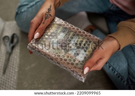 Partial view of hands of girl holding and packing family photo in air bubble film on blurred floor. Young woman with tattoos moving to new apartment. Concept of home relocation