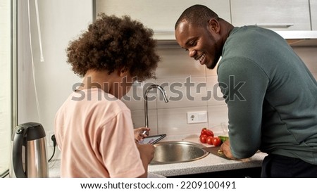 Smiling black man cook salad and look at little son draw humans on digital tablet at home kitchen. Family lifestyle and relationship. Fatherhood and parenting. Domestic leisure and entertainment