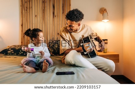 Happy single dad smiling at his daughter while playing a guitar at home. Cheerful dad and daughter having a good time while sitting on a bed together. Father and daughter spending some quality time. Royalty-Free Stock Photo #2209103157