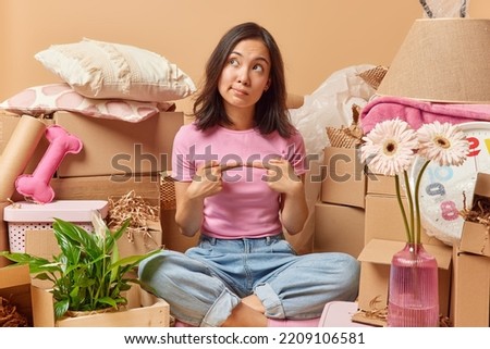 Thoughtful Asian woman sits crossed legs keeps index fimgers opposite each other thinks about relocation and changing place of living dressed in casual clothes surrpunded by cardboard boxes. Moving