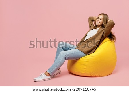 Full body young successful employee business woman 30s she wear casual brown classic jacket sit in bag chair look camera hold hands behind neck isolated on plain pastel light pink background studio