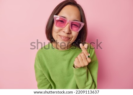 Lovely young Asian woman with dark hair makes korean like sign expresses love shapes mini heart wears trendy sunglasses and green pullover isolated over pink studio background likes something