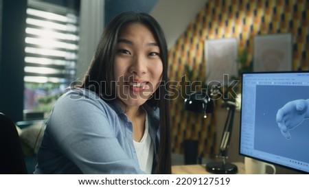 Surprised asian woman rejoicing and looking at camera while sitting at the table at home office and working remotely on 3D modeling project on computer